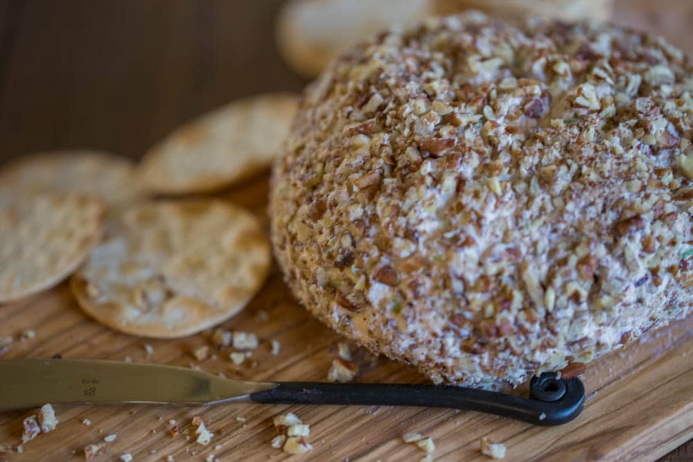 A cheese ball on a wood cutting board next to a knife with crackers in the background