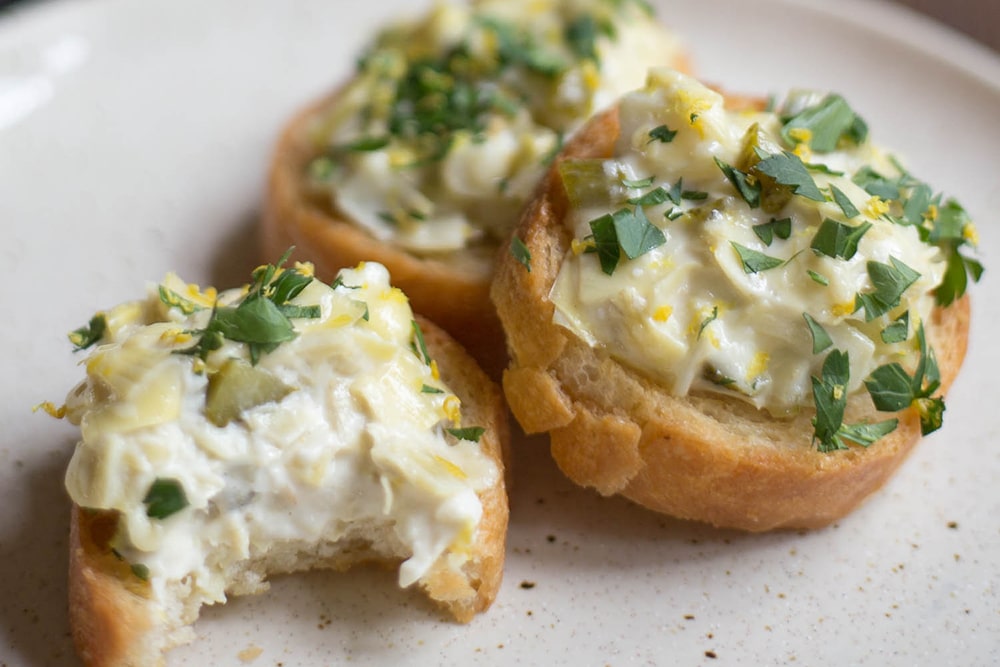 A plate with 3 crostini topped with artichoke spread garnished with chopped parsley