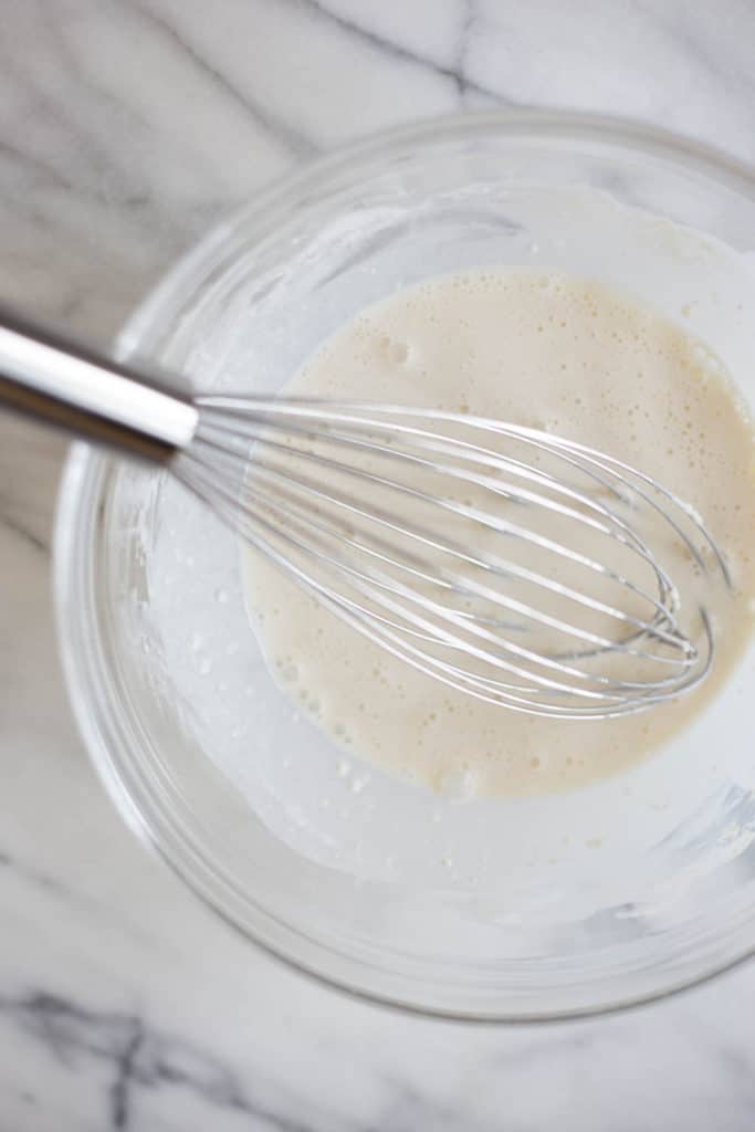 An overhead view of a clear bowl filled with a mixture of half and half and flour with a whisk