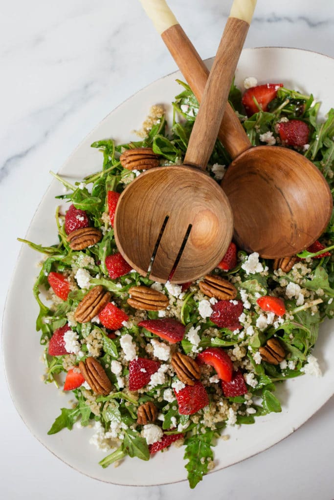 Overhead view of a white platter of salad with strawberries, goat cheese, and pecans with wooden serving spoons
