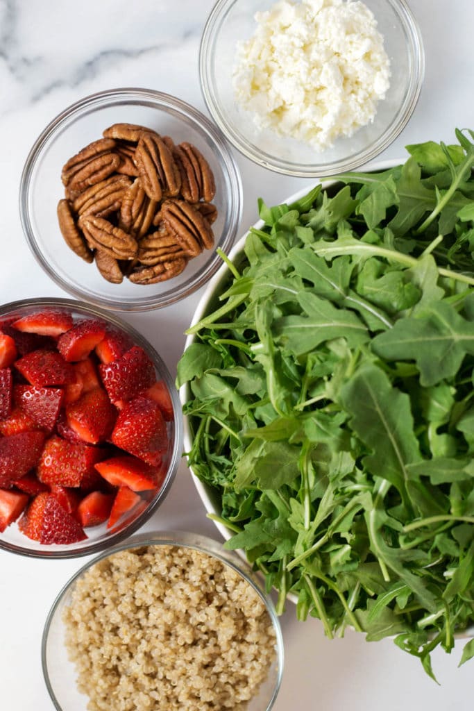 Overhead view of a bowl of arugula, a bowl of quinoa, a bowl of chopped strawberries, a bowl of pecans, and a bowl of crumbled goat cheese