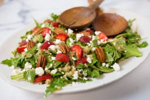 a white platter of salad with strawberries, goat cheese, and pecans with wooden serving spoons