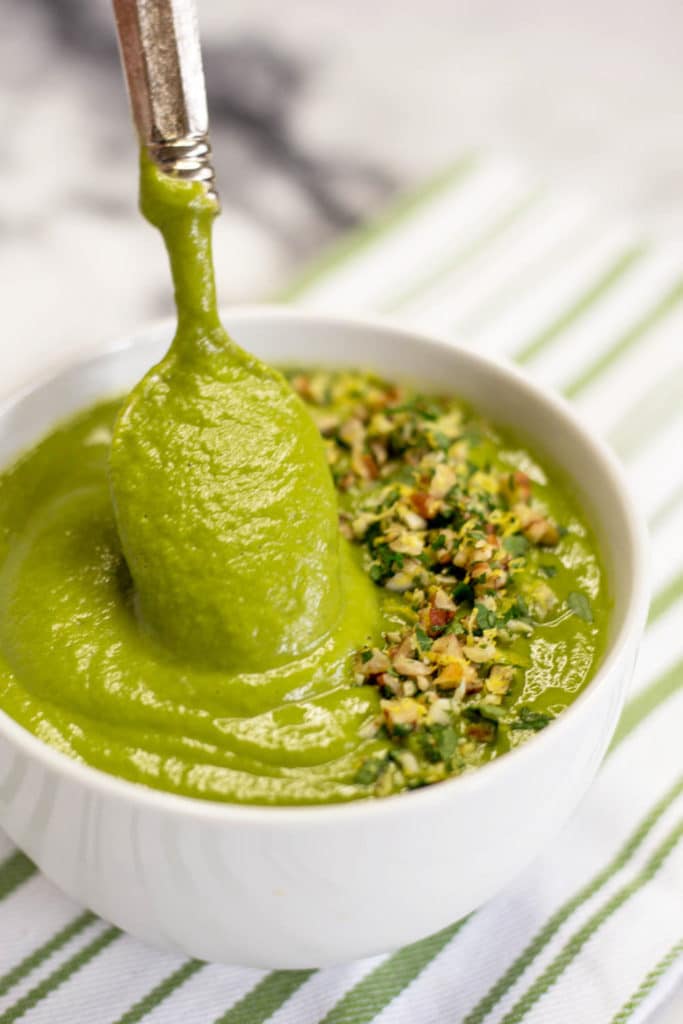 A spoon dipped into a white bowl of spinach soup garnished with pecan gremolata