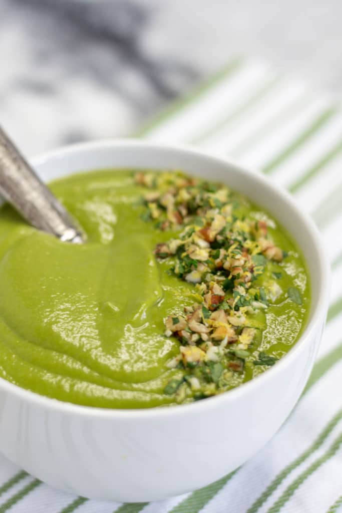 A white bowl of spinach soup garnished with pecan gremolata on a green striped napkin
