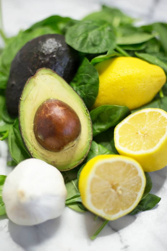 A bulb of garlic, a lemon cut in half, an avocado cut in half with the pit exposed, a whole lemon, on a bed of fresh spinach