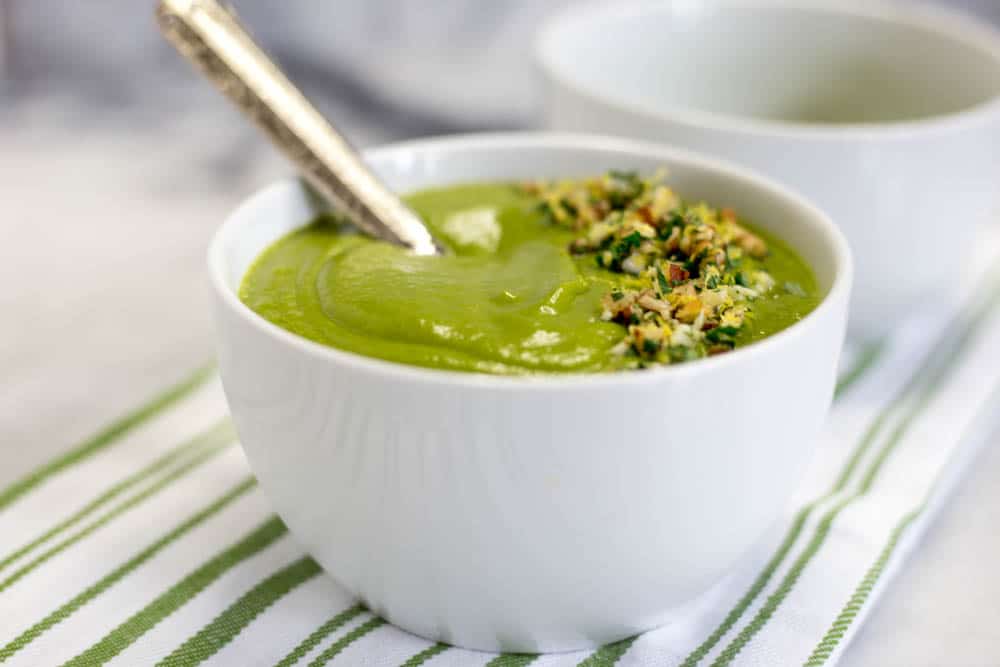 A white bowl of spinach soup garnished with pecan gremolata and a spoon