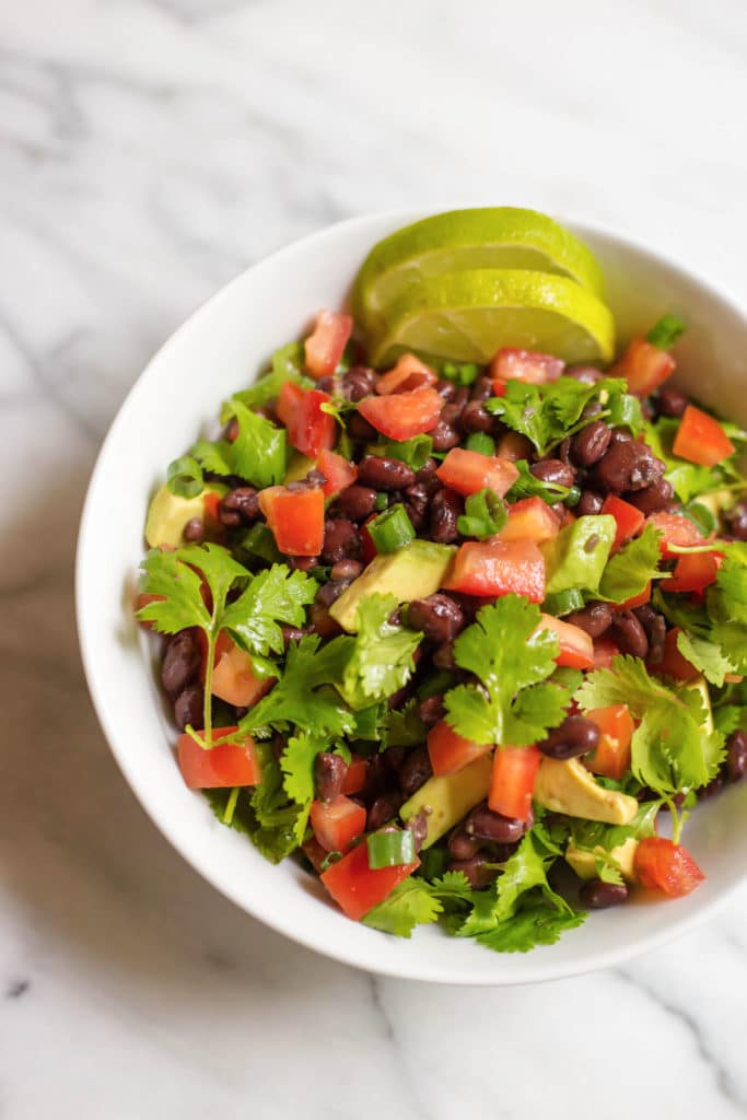 White bowl of salad with chopped tomatoes, chopped avocado, cilantro, black beans, and 2 slices of lime
