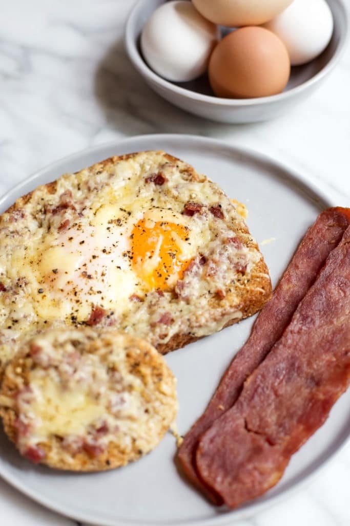 A piece of bread with a sunny side up egg in the center topped with cheese, bacon, and a bowl of fresh eggs