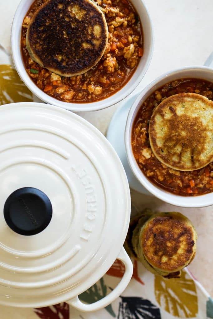 Overhead shot of white dutch oven with lid and two white bowls of buffalo chicken chili topped with pancakes