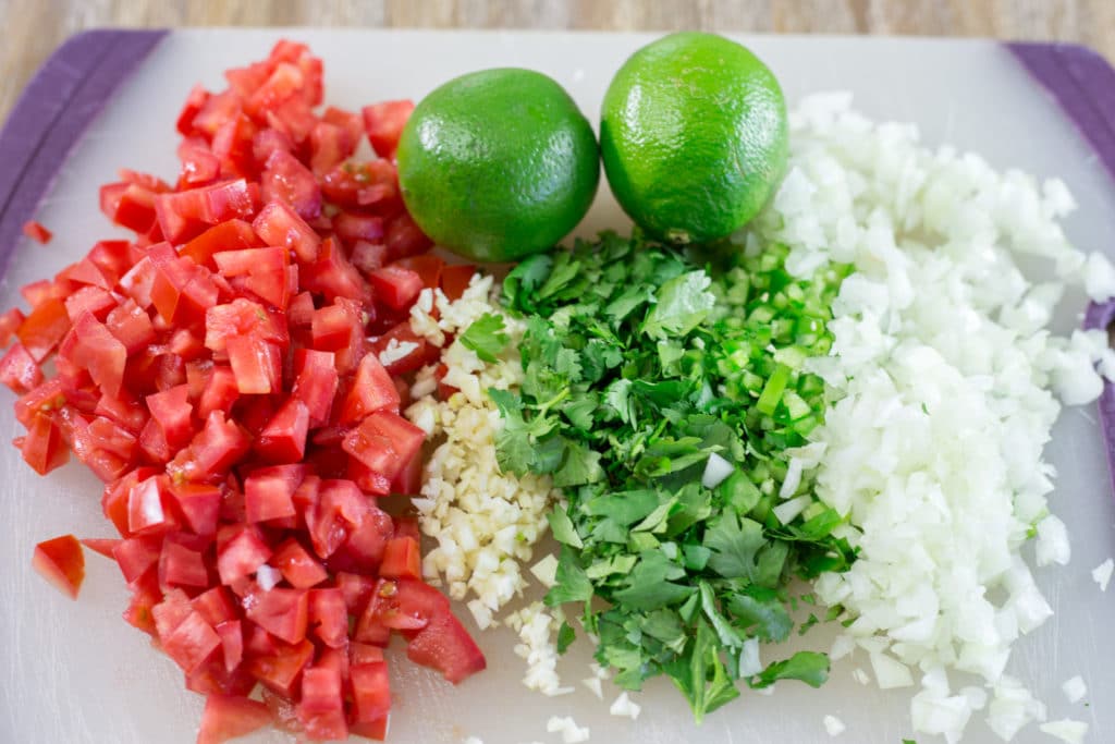 Diced tomatoes, chopped garlic, chopped cilantro, diced onions, and 2 limes on a cutting board
