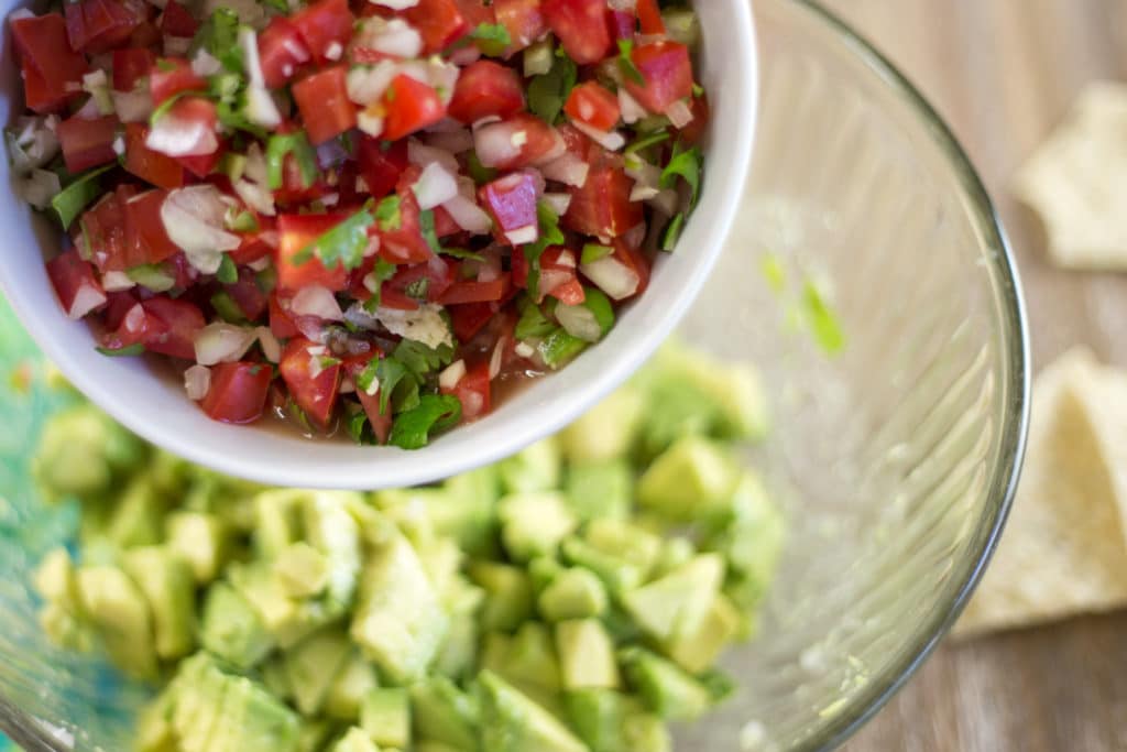 A white bowl of pico de gallo being added to a clear bowl of diced avocado