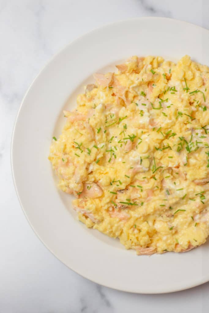 White plate of scrambled eggs with salmon and cream cheese garnished with chopped chives