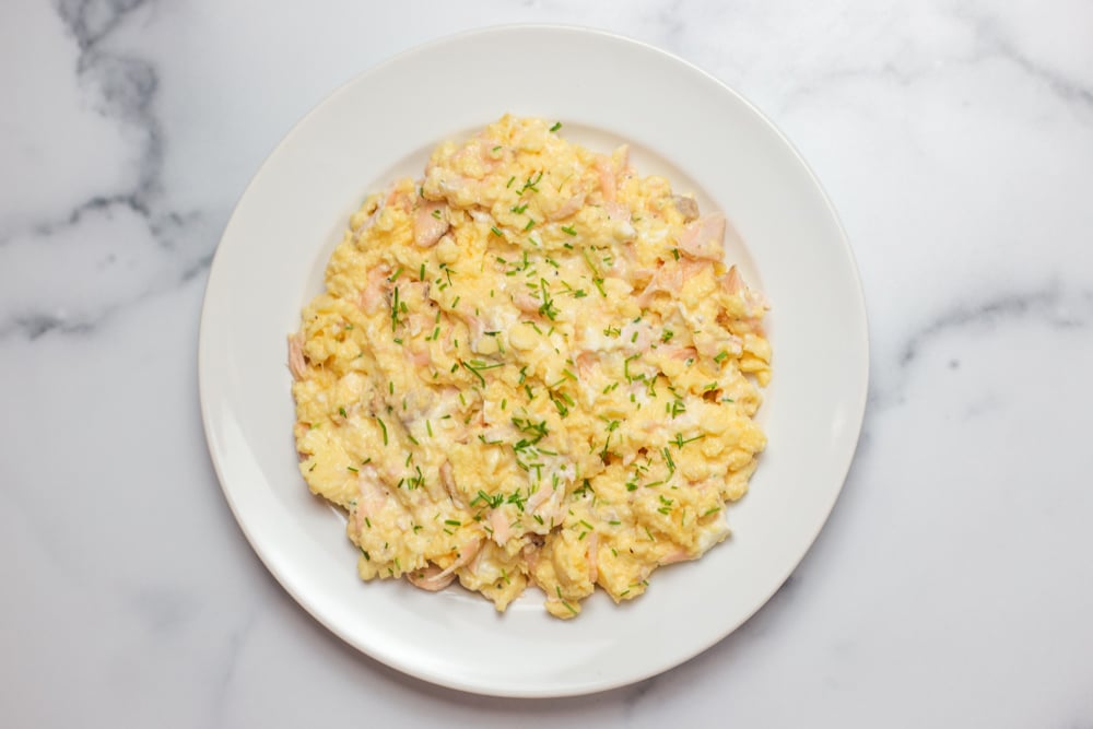 White plate of scrambled eggs with salmon and cream cheese garnished with chopped chives