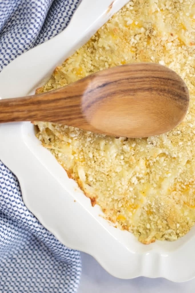 Overhead view of a white baking dish with hash brown casserole topped with a wooden spoon and a blue checked napkin on the side