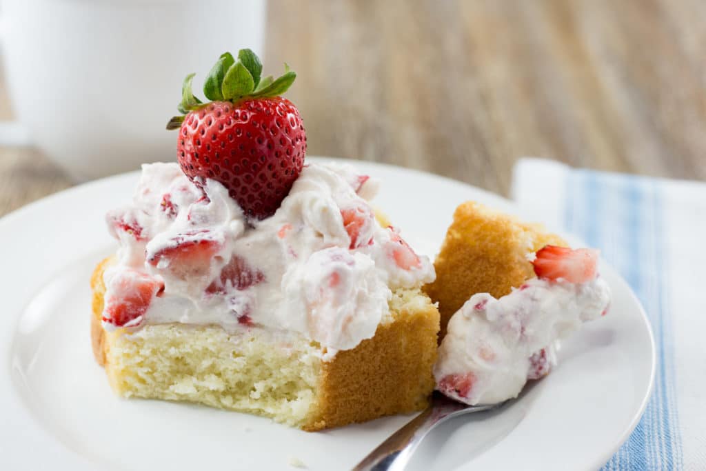 White plate with a piece of pound cake topped with whipped cream and a strawberry with a bite taken out