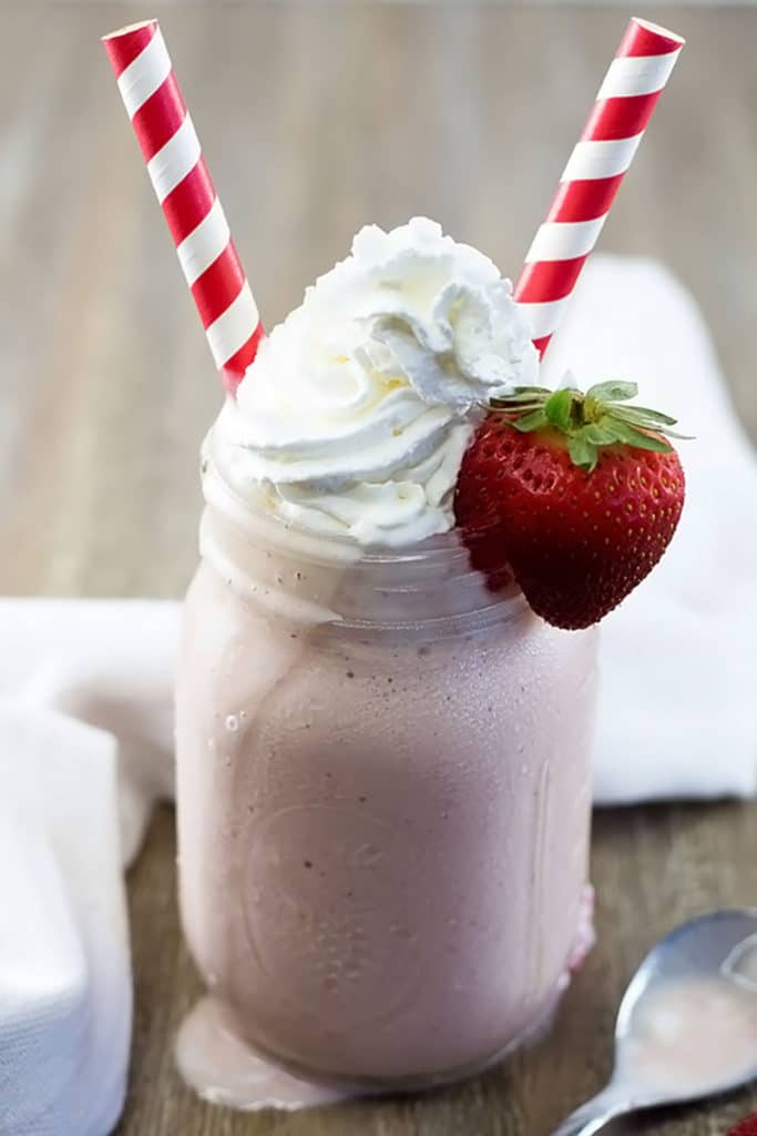 A mason jar filled with strawberry milkshake topped with whipped cream, a strawberry on the edge, and 2 red striped straws