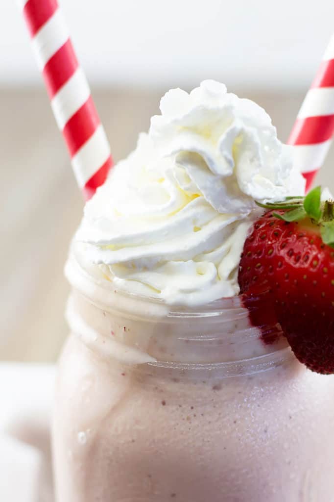 Up close view of a mason jar filled with strawberry milkshake topped with whipped cream, a strawberry on the edge, and 2 red striped straws