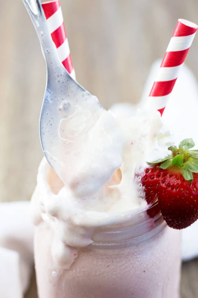 A spoon scooping melted whipped cream out of a mason jar filled with strawberry milkshake with a strawberry on the edge and 2 red striped straws