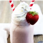 A mason jar filled with strawberry milkshake topped with whipped cream with a strawberry on the edge and 2 red striped straws