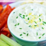 A bowl of creamy white dip garnished with chives and lemon zest surrrounded by celery sticks and red pepper sticks