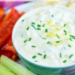A bowl of creamy white dip garnished with lemon zest and chopped chives surrounded by celery sticks and red pepper sticks