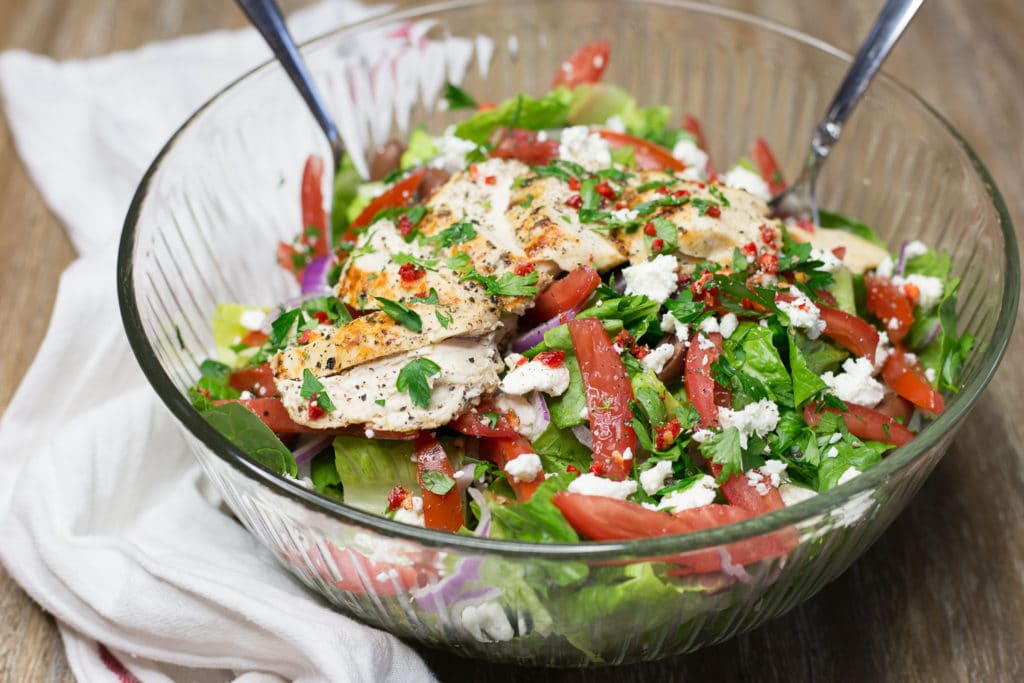 A clear bowl of salad with tomatoes, feta cheese, and sliced chicken