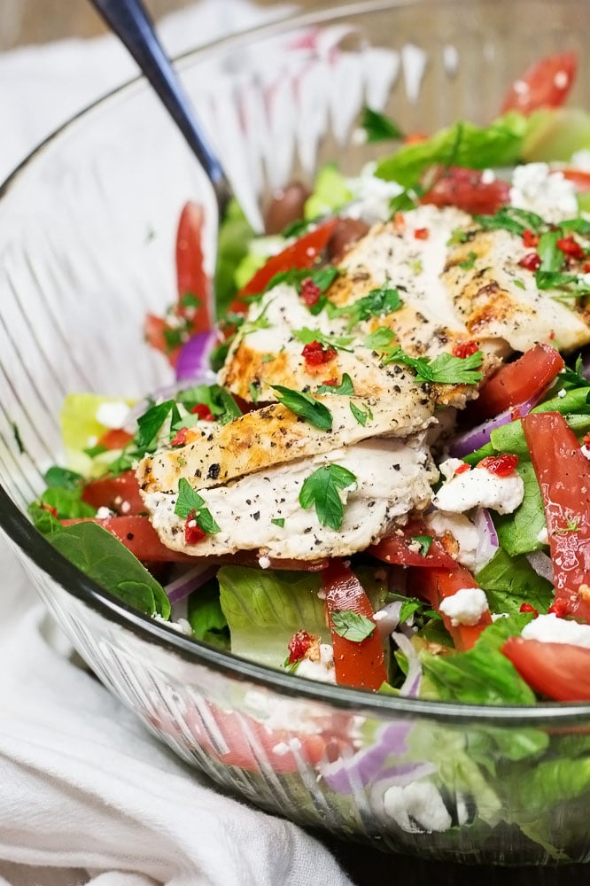 A clear bowl of salad with tomatoes, feta cheese, parsley, and slices of chicken breast