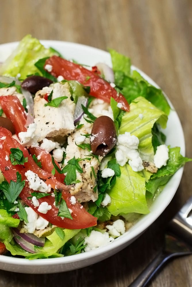 A white bowl of salad with tomatoes, chunks of chicken, olives, and feta cheese