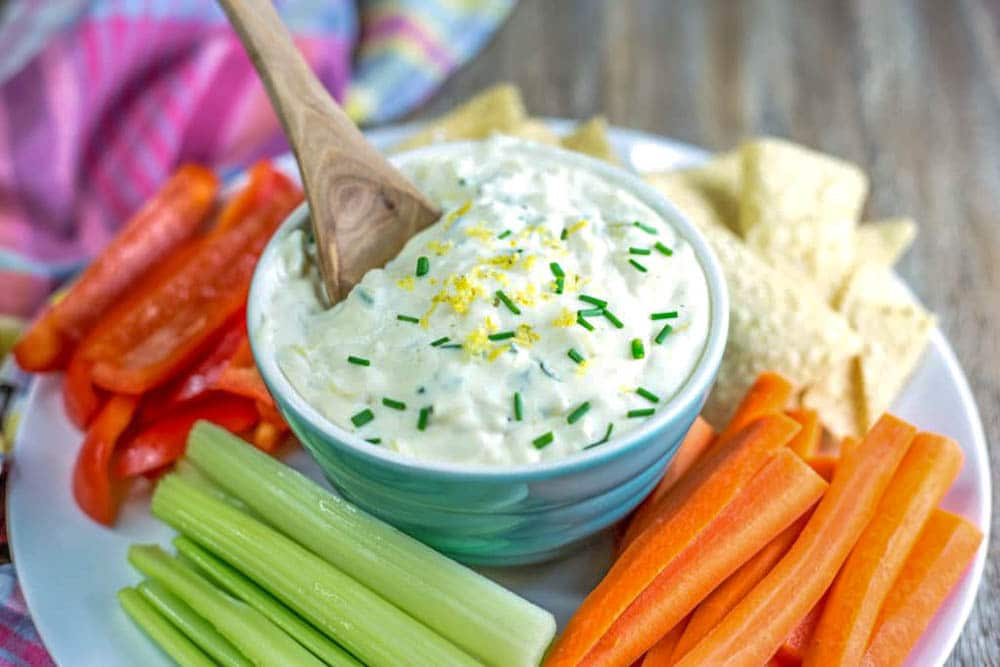A bowl of white dip garnished with chives and lemon zest surrounded by celery, carrots, and red pepper sticks