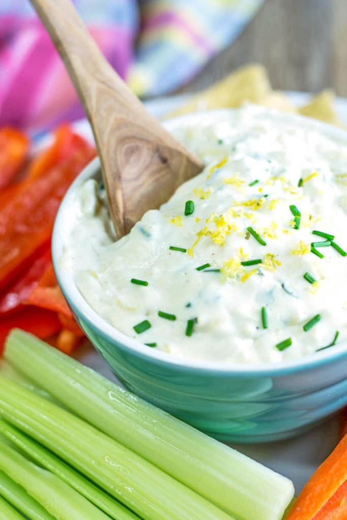 A bowl of white dip garnished with chives and lemon zest surrounded by celery, carrots, and red pepper sticks