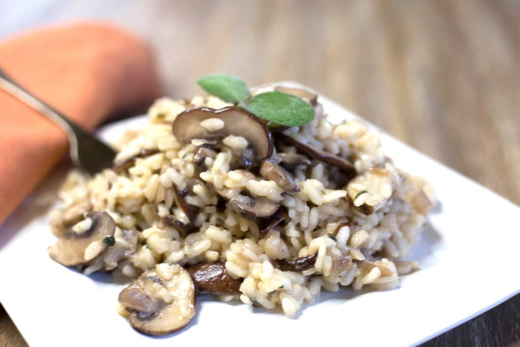 A white plate of mushroom risotto garnished with sage next to a burnt orange napkin