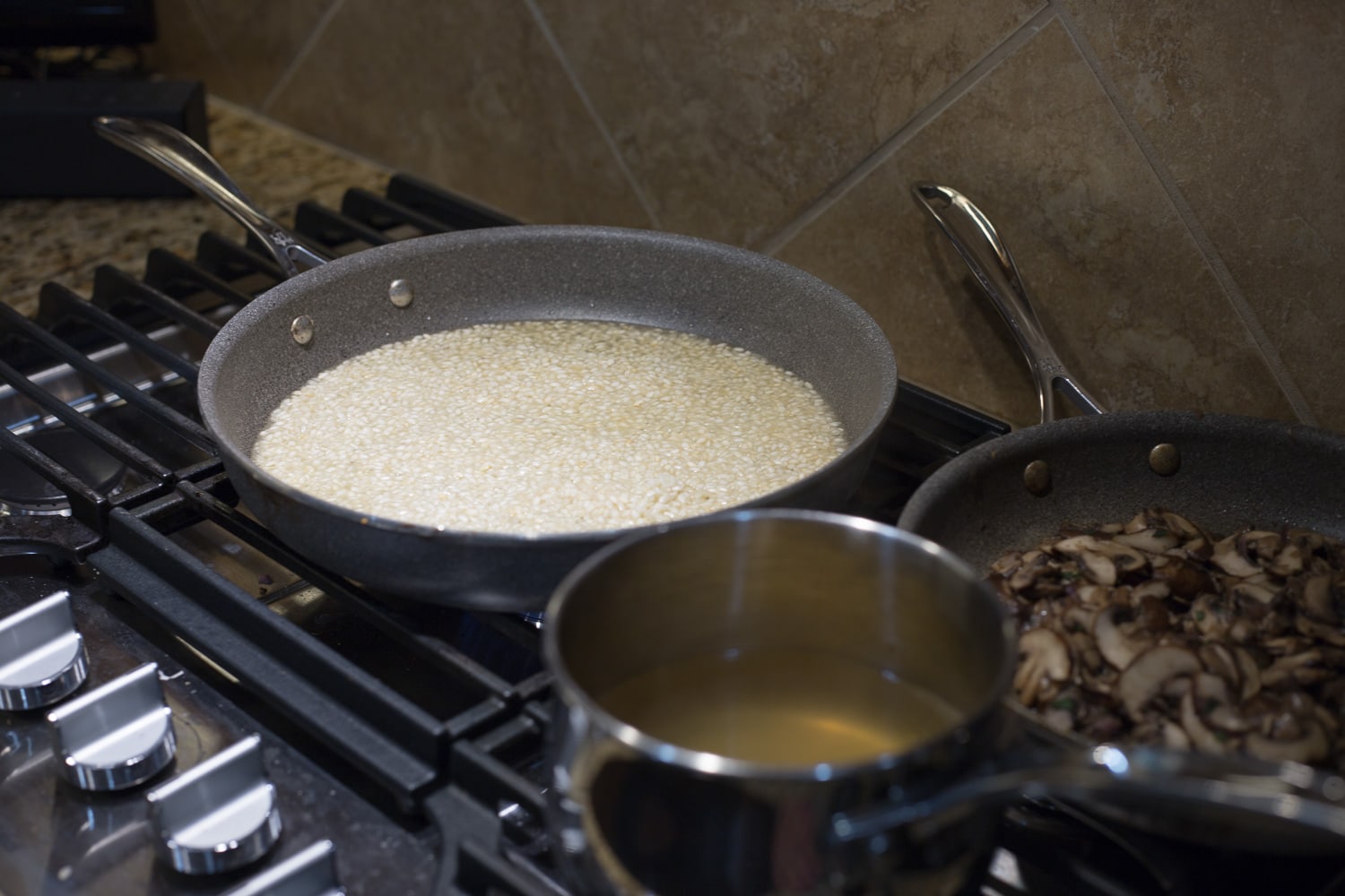 A gas cooktop with a saucepan of chicken broth, a skillet of partially cooked arborio rice, and a skillet of cooked mushrooms