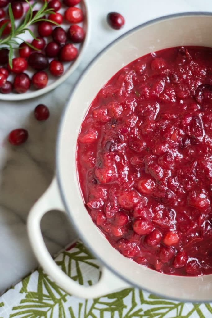 Overhead view of a white dutch oven filled with bright red cranberry sauce and a white plate of fresh cranberries