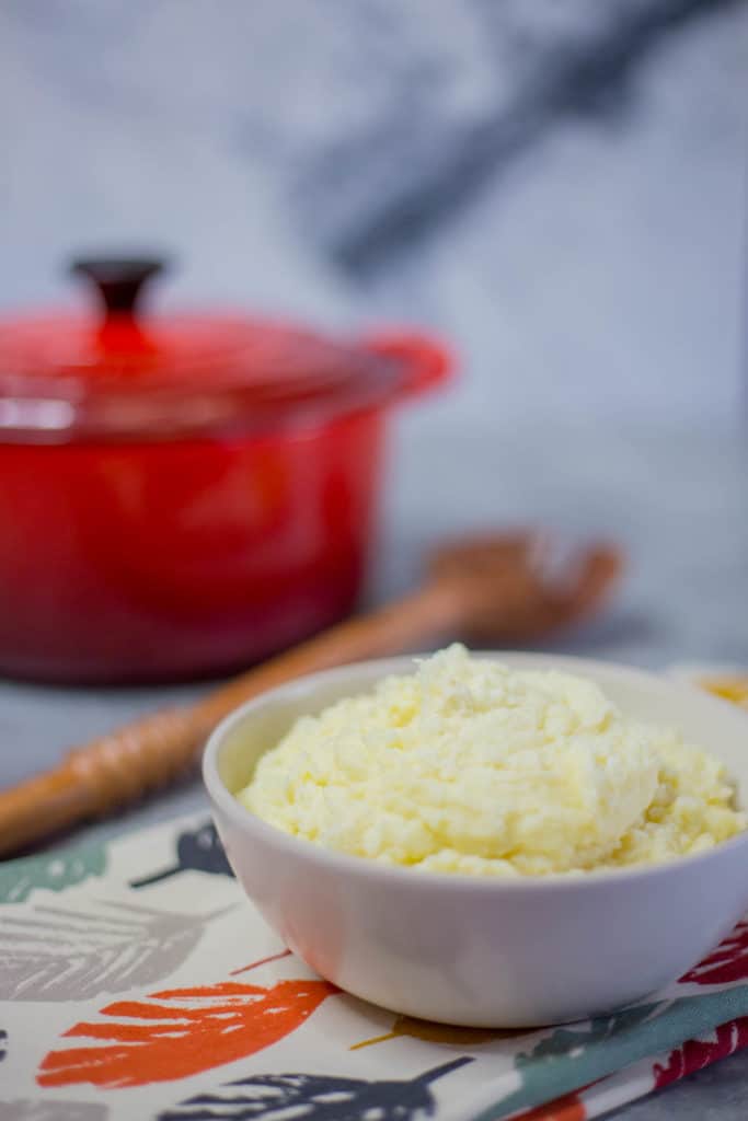 A white bowl filled with mashed potatoes on a fall colored napkin with a wooden spoon and a red dutch oven blurred in the background