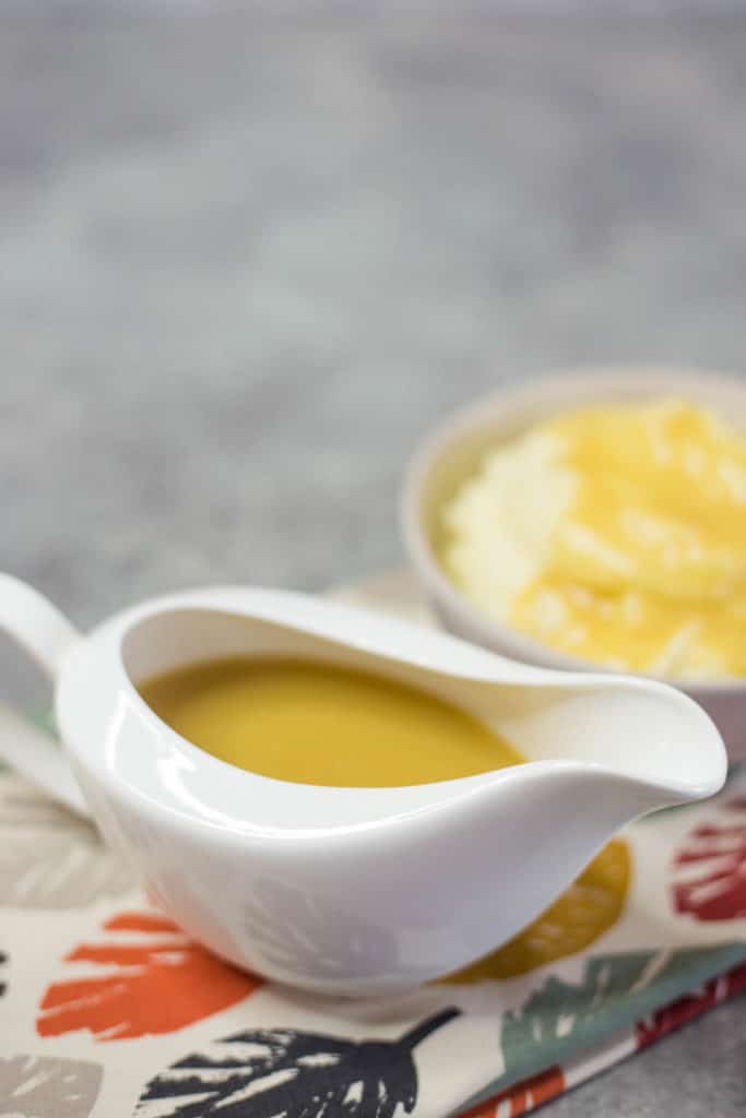 A white gravy boat filled with gravy on a napkin next to a bowl of mashed potatoes and gravy