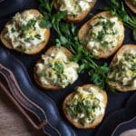 A black tray topped with small round crostini with a creamy topping garnished with chopped parsley
