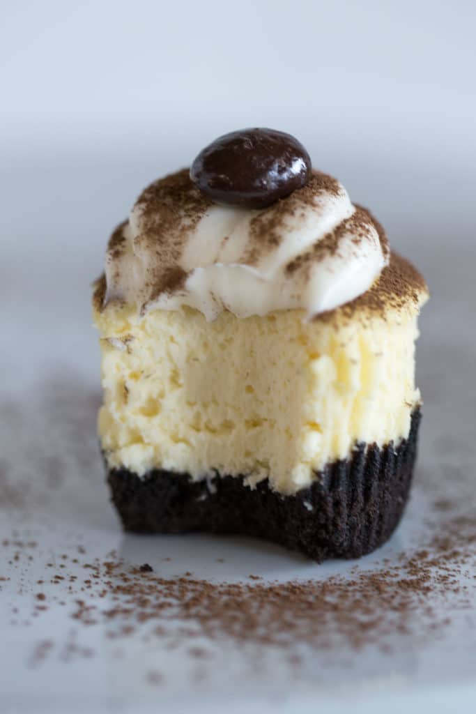 Up close view of a cheesecake cupcake with a bite out of it topped with white frosting, cocoa powder, and an espresso bean