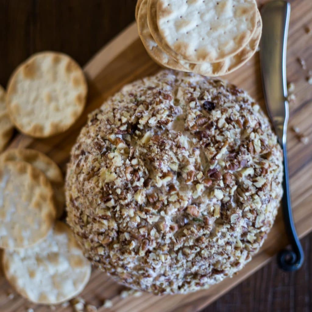 Overhead view of a cheese ball coated in pecan pieces on a wood cutting board next to a knife surrounded by crackers
