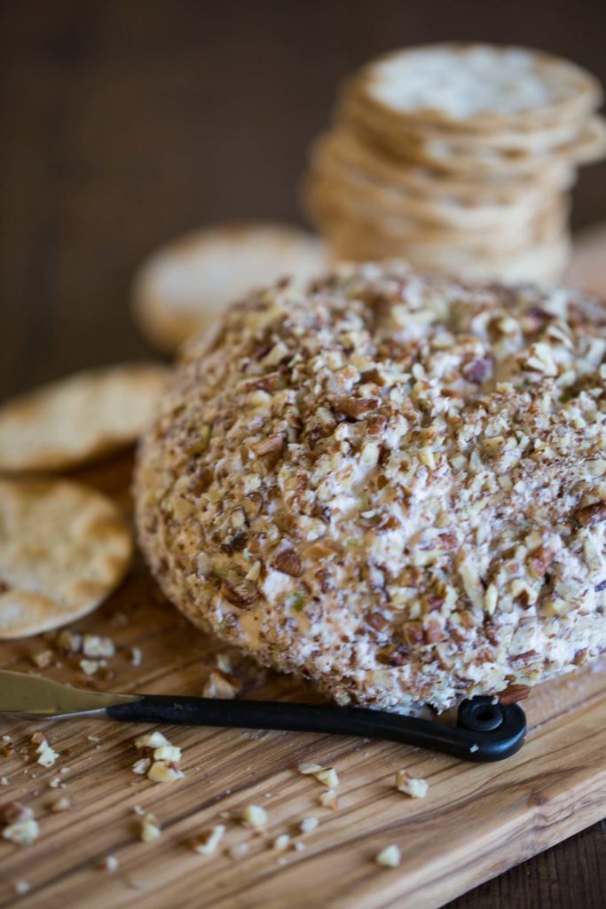 A cheese ball coated with pecan pieces on a wood cutting board with crackers in the background