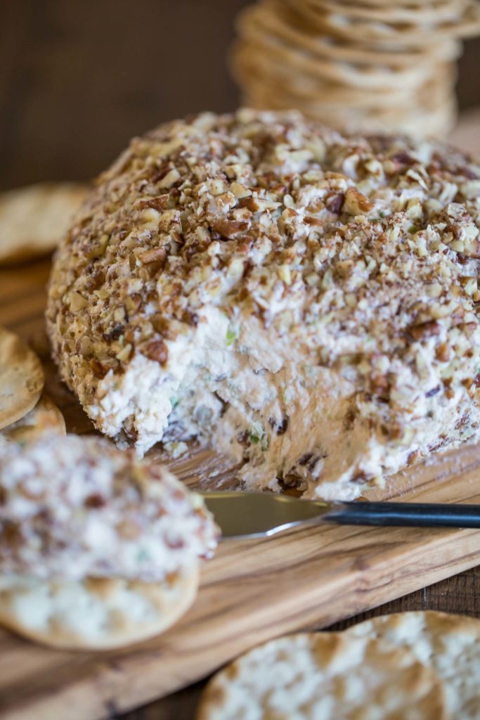 A cheese ball coated in pecan pieces on a wood cutting board with a portion removed 