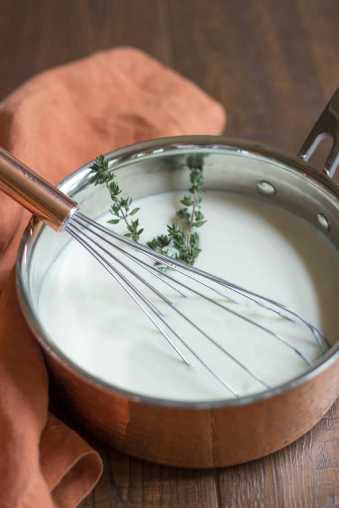A copper saucepan full of bechamel sauce with a whisk and a sprig to thyme