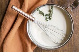 Overhead shot of saucepan with bechamel, a whisk, and a sprig of thyme