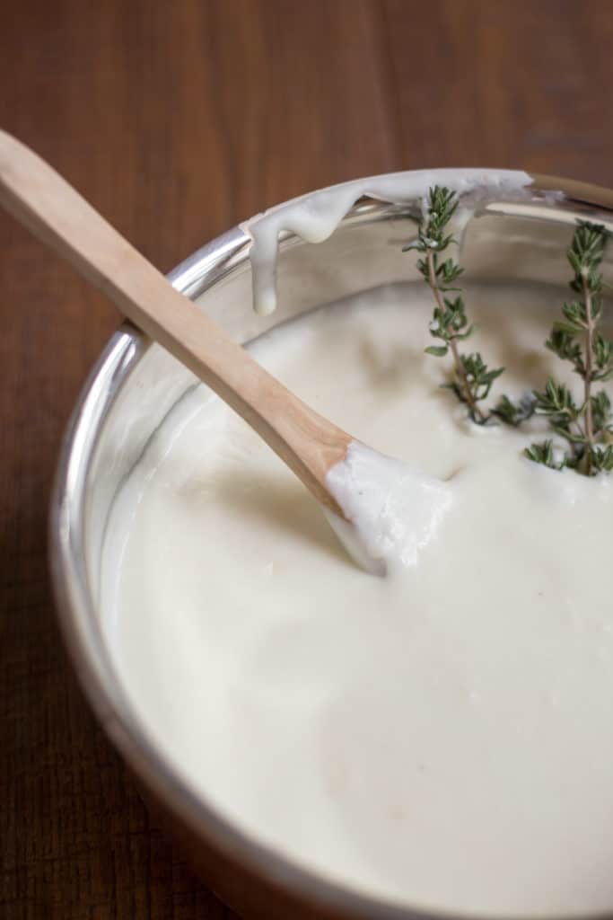 A saucepan of bechamel sauce with a wooden spoon and a sprig of thyme