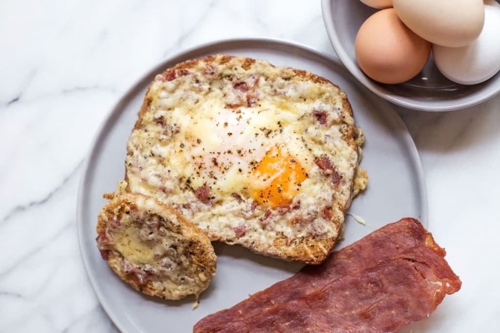 A piece of bread with a sunny side up egg in the center, bacon, and a bowl of fresh eggs