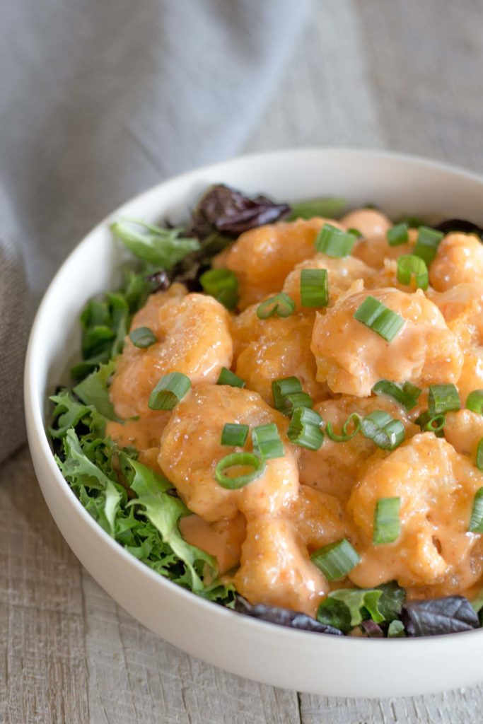 White bowl of firecracker shrimp over a bed of greens with a green onion garnish
