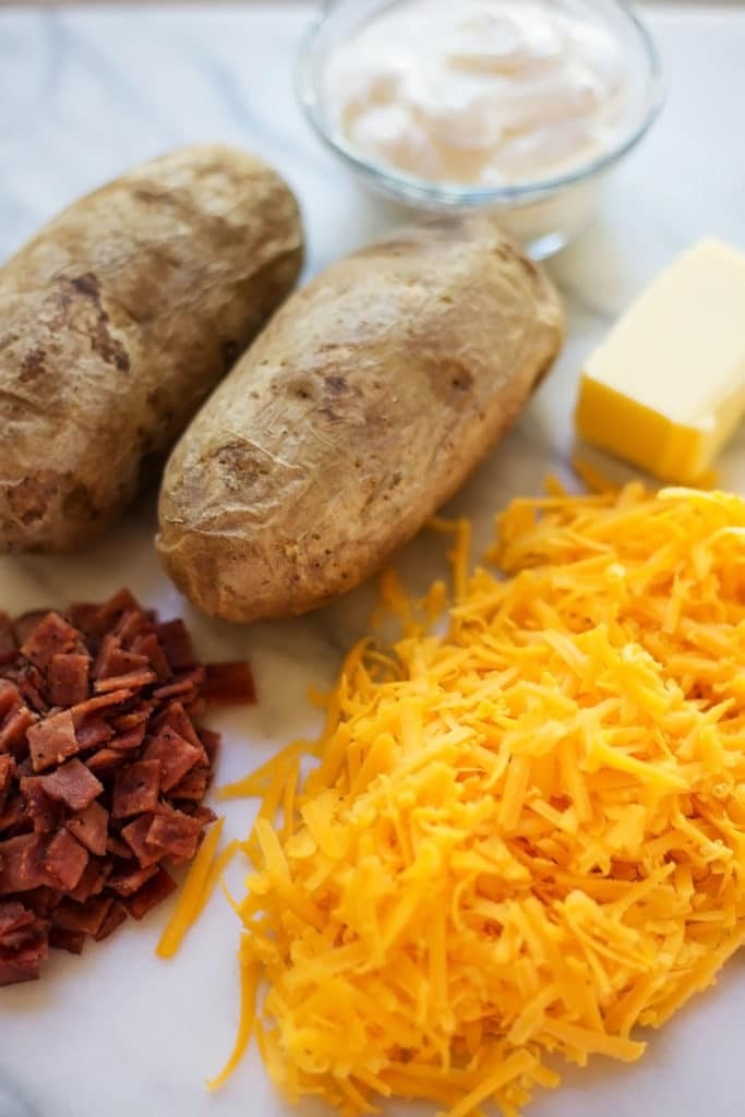 A mound of chopped cooked bacon, a mound of shredded cheddar cheese, two baked potatoes, a pad of butter, and a bowl of sour cream