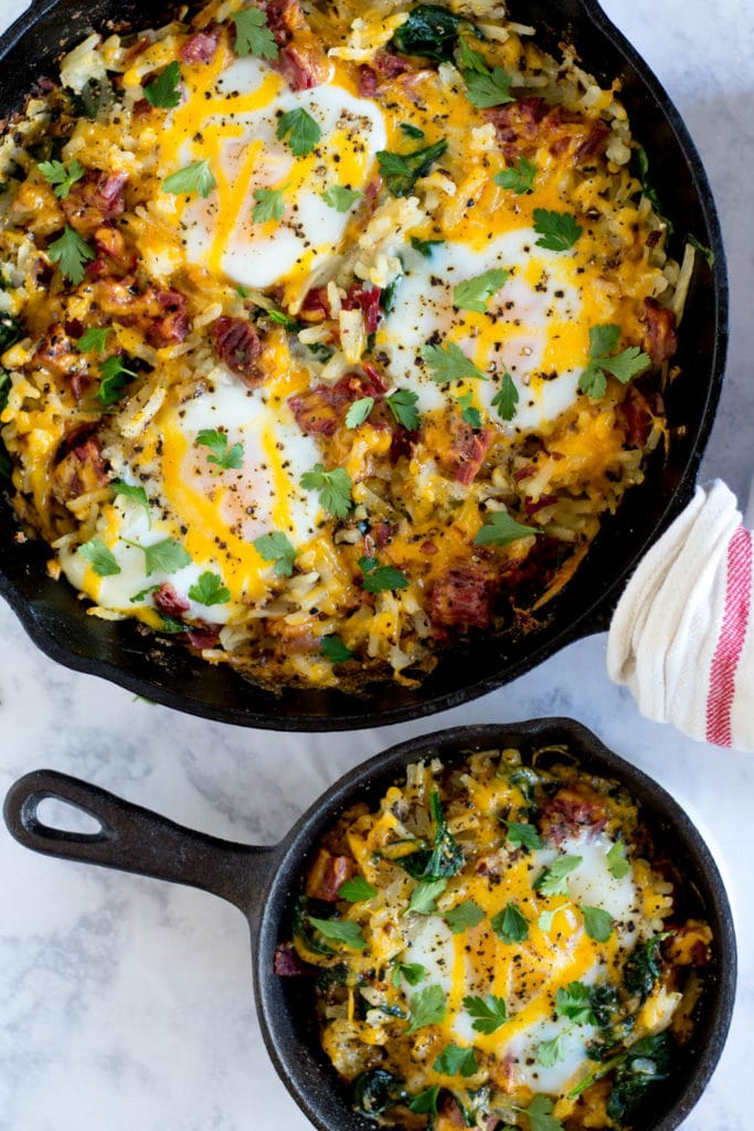 Large skillet of corned beef has with baked eggs next to small skillet of corned beef hash