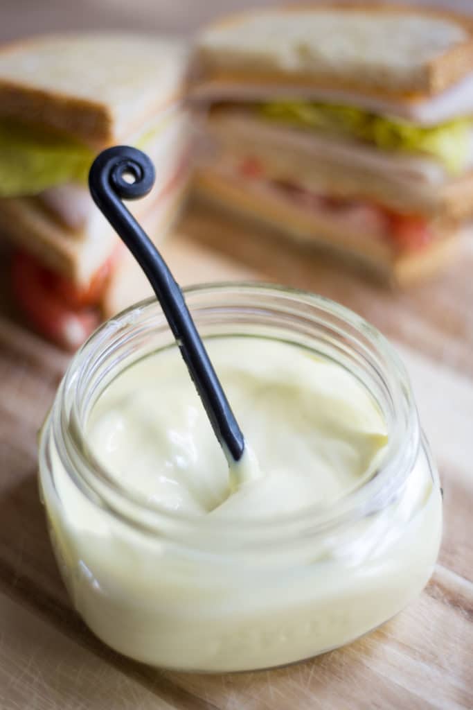 A spoon in a clear jar of homemade mayonnaise with a sandwich cut in half in the background