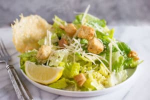 A white bowl of caesar salad with croutons, parmesan cheese, a lemon slice, and a parmesan cheese crisp
