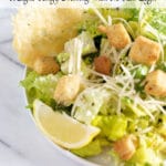 A white bowl of caesar salad with croutons, parmesan cheese, a lemon slice, and a parmesan cheese crisp